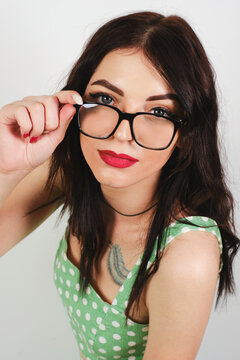 A young pretty girl in glasses looks frowningly at the camera on a light background in a polka-dot dress.