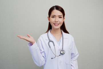 An Asian female doctor puts her hand to the side to make a presentation. with a smile, put the headphones on her shoulder wearing a white coat.