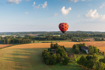 Red hot air balloon with black dots flying over fields and forests on a sunny summer morning. Aerial view, Latvia.