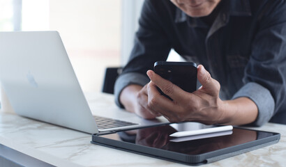 Close up asian business man in casual wear using mobile smart phone during working on laptop computer at modern home office with diigtal tablet on table