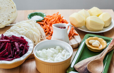 Ingredients for soup cooking in multi-colored bowls on a wooden cutting board - onions, carrots, beets, garlic, cabbage, salt, tomato paste, potatoes.