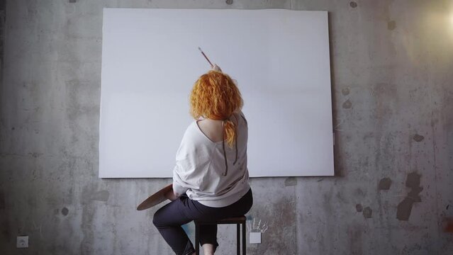 Female painter sitting in front of big white canvas holding paintbrushes and palette. Caucasian redhead artist waiting for inspiration meditating in front of blank canvas. High quality 4k footage