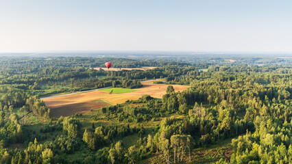Fototapeta na wymiar Red hot air balloon with black dots flying over fields and forests on a sunny summer morning. Aerial view, Latvia.
