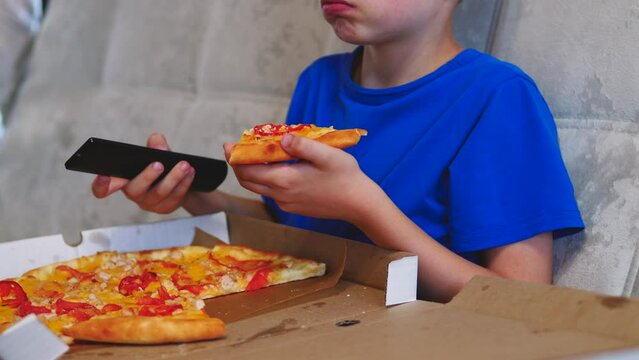 caucasian boy 8 years old eats pizza and watches tv holding the remote control in his hand while sitting on the couch. The child watches cartoons and eats fast food. Relax watching TV