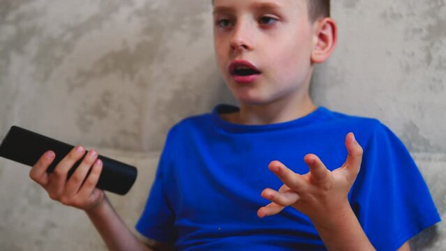  cute Caucasian boy 6-7 years old on the couch with a pizza in his hand and a TV remote control is looking for an interesting program. The child drops a piece of pizza from his hands from what he sa