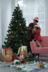 Happy latino family, young bearded father wear santa hat and little daughter decorating a Christmas tree together with star, gift box, pine cones, balls and candies at living room at home