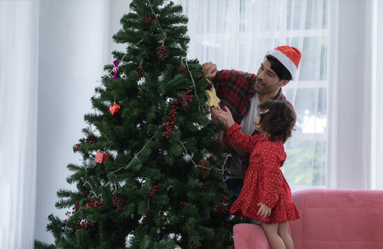 Happy Latino Family, Young Bearded Father Wear Santa Hat And Little Daughter Decorating A Christmas Tree Together With Star, Gift Box, Pine Cones And Candies At Living Room At Home