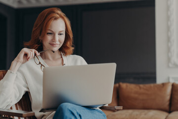 Fototapeta na wymiar Horizontal shot of happy skilled ginger woman teacher works distantly, checks students works via laptop, holds spectacles, poses in modern cozy apartment. Student checks database for university