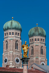 Die goldene Statue der Mariensäule auf dem Marienplatz in der Münchner Altstadt, eingerahmt von den zwei Türmen der Frauenkirche bei sonnigem Wetter und wolkenlosem, blauen Himmel