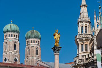 Fototapeta na wymiar Die vergoldete Statue von Maria mit Jesus auf dem Arm auf der Mariensäule auf dem Marienplatz in der Altstadt von München eingerahmt von den beiden Türmen der Frauenkirche und einem Zierturm des Neuen