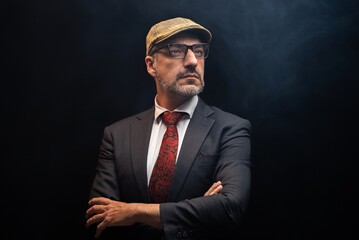 Portrait of detective in elegant black suit and red tie wearing eye glasses and hat standing with crossed arms. isolated on black background.