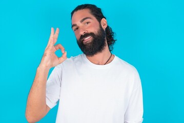 young bearded man wearing white T-shirt over blue studio background hold hand arm okey symbol toothy approve advising novelty news