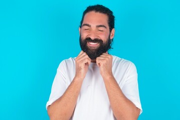 young bearded man wearing white T-shirt over blue studio background grins joyfully, imagines something pleasant, copy space. Pleasant emotions concept.