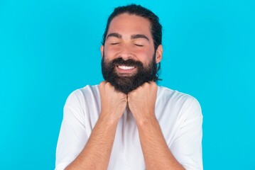 Cheerful young bearded man wearing white T-shirt over blue studio background has shy satisfied expression, smiles broadly, shows white teeth, People emotions