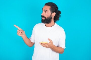 Fototapeta premium Stunned young bearded man wearing white T-shirt over blue studio background points sideways right copy space, recommends product, sees astonishing thing