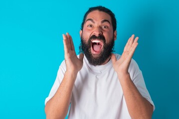 Surprised happy young bearded man wearing white T-shirt over blue studio background , glad to see big discounts on clothes, expresses shock, keeps hands near head, jaw dropped.