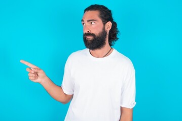 young bearded man wearing white T-shirt over blue studio background points at copy space and advertises something, advices best price.