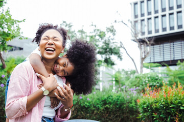 Playful little girl with curly dark hair is hugging her mum's neck and looking at her with a smile, they have perfect weekends at park. Good family relations concept
