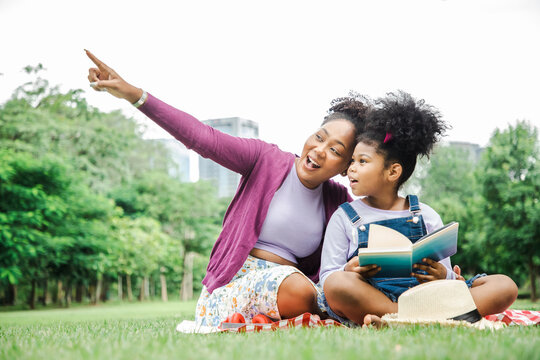 Happy African American young family mother and child little girl having fun and enjoying outdoor laying on picnic blanket reading book at summer garden spring park, Family relaxation concept