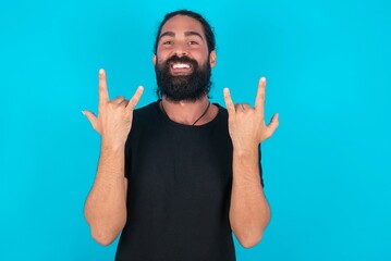 young bearded man wearing black T-shirt over blue studio background makes rock n roll sign looks...