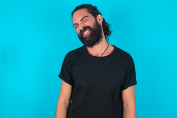 Positive young bearded man wearing black T-shirt over blue studio background with overjoyed expression closes eyes and laughs shows white perfect teeth