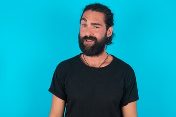 Portrait of dissatisfied young bearded man wearing black T-shirt over blue studio background smirks face, purses lips and looks with annoyance at camera, discontent hearing something unpleasant