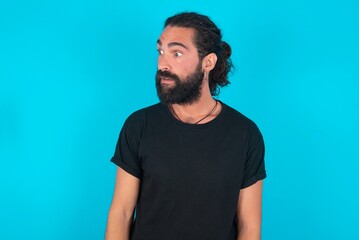 Fototapeta premium young bearded man wearing black T-shirt over blue studio background stares aside with wondered expression has speechless expression. Embarrassed model looks in surprise