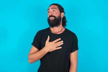 Joyful young bearded man wearing black T-shirt over blue studio background expresses positive...