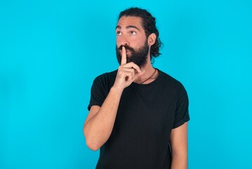 young bearded man wearing black T-shirt over blue studio background silence gesture keeps index finger to lips makes hush sign. Asks not to share secret