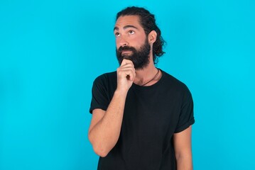 Fototapeta premium Thoughtful young bearded man wearing black T-shirt over blue studio background holds chin and looks away pensively makes up great plan