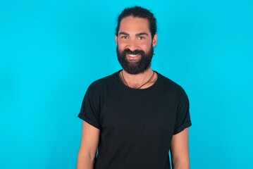 young bearded man wearing black T-shirt over blue studio background  with nice beaming smile pleased expression. Positive emotions concept