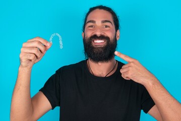 young bearded man wearing black T-shirt over blue studio background holding an invisible aligner and pointing perfect straight teeth. Dental healthcare and confidence concept.