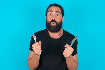 hungry young bearded man wearing black T-shirt over blue studio background holding in hand fork...