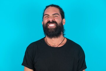 Fototapeta premium young bearded man wearing black T-shirt over blue studio background keeps teeth clenched, frowns face in dissatisfaction, irritated because of much duties.