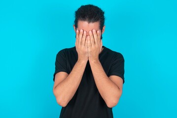 young bearded man wearing black T-shirt over blue studio background covering her face with her...