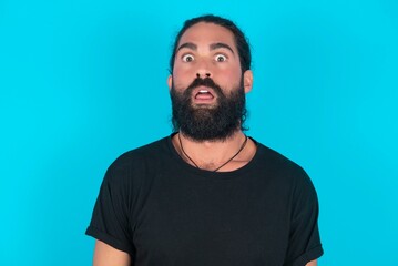 Oh my God. Surprised young bearded man wearing black T-shirt over blue studio background stares at camera with shocked expression exclaims with unexpectedness,