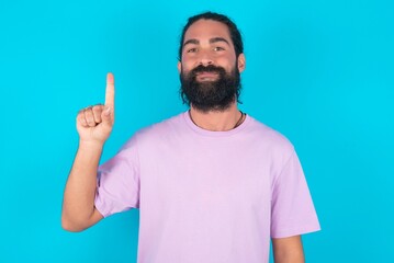 young bearded man wearing violet T-shirt over blue studio background showing and pointing up with finger number one while smiling confident and happy.