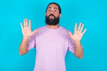 Naklejka premium young bearded man wearing violet T-shirt over blue studio background Moving away hands palms showing refusal and denial with afraid and disgusting expression. Stop and forbidden.