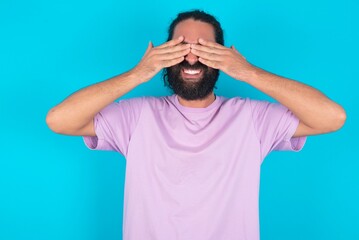young bearded man wearing violet T-shirt over blue studio background covering eyes with hands smiling cheerful and funny. Blind concept.