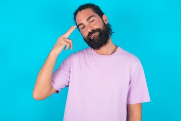 Fototapeta premium Unhappy young bearded man wearing violet T-shirt over blue studio background makes suicide gesture and imitates gun with hand, curves lips, keeps two fingers on temple