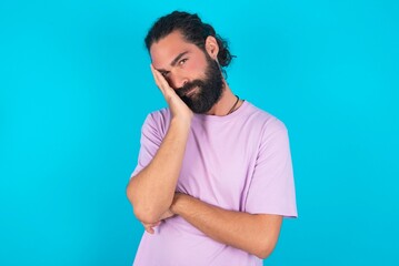 Very bored young bearded man wearing violet T-shirt over blue studio background holding hand on cheek while support it with another crossed hand, looking tired and sick,