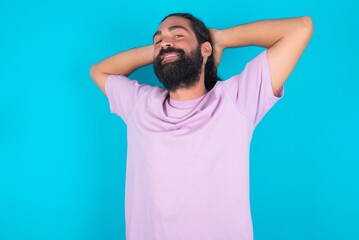 young bearded man wearing violet T-shirt over blue studio background stretching arms, relaxed position.