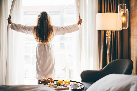 Young woman in bathrobe opening the curtains in hotel room.