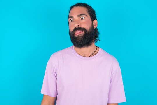 young bearded man wearing violet T-shirt over blue studio background showing grimace face crossing eyes and showing tongue. Being funny and crazy