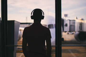 Fototapeta na wymiar Silhouette of man with headphones while waiting for flight. Traveler looking from window of airport terminal before boarding to airplane..
