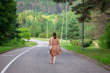 a woman in a dress with a handbag on her shoulder and a braided ponytail walking down an empty road barefoot, carrying her shoes in her hands