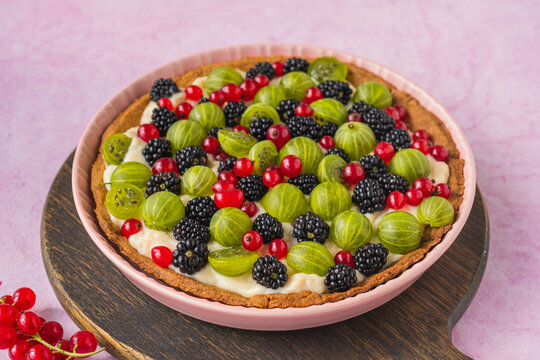 Dessert, shortbread tart with assorted berries and custard in a pink ceramic form on a pink concrete background. Dessert recipes with berries.