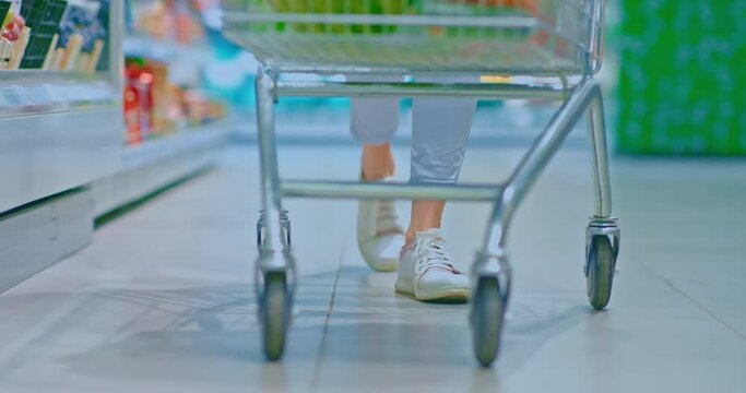 Close-up, women's feet in sneakers walking through the supermarket. Woman is driving a grocery cart. Shopping to the supermarket for groceries. 4k, ProRes