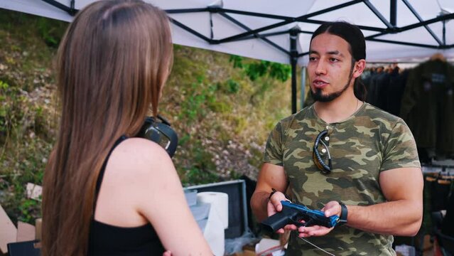 White bearded man in camo t-shirt holding handgun showing it to female client interested in buying it. Buying and selling firearms topic. Outdoor horizontal shot. High quality 4k footage