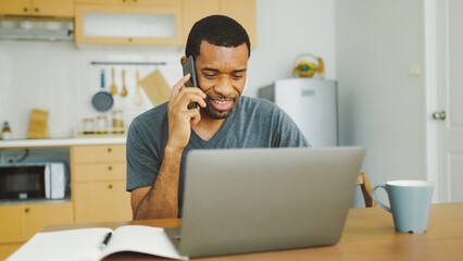 Happy Smiling Black African American man talking on mobile phone while working on laptop computer at home.